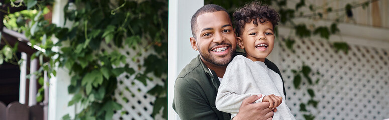 happy african american father in braces hugging cute curly son and sitting on porch on house, banner