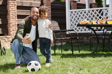 happy african american father and son holding smartphone while standing on backyard of house