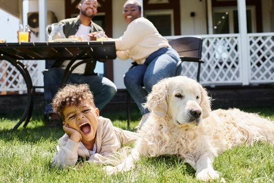 emotional african american boy lying on green lawn near dog and parents having lunch in garden