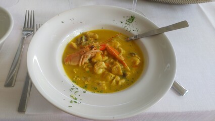On a table covered with a white tablecloth are a ceramic plate with seafood soup and greens. A metal fork rests next to it. Dinner time