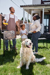 cheerful african american man cooking grilled corn on bbq grill near dog, wife and son on backyard