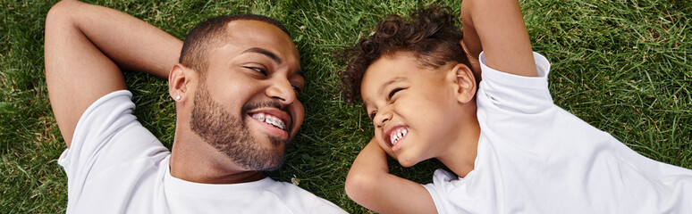 top view of cheerful african american father and son lying on green lawn on backyard, family banner - 740868722