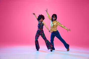 Man and woman dressed in vibrant 1970s attire dancing in motion against gradient pink studio...