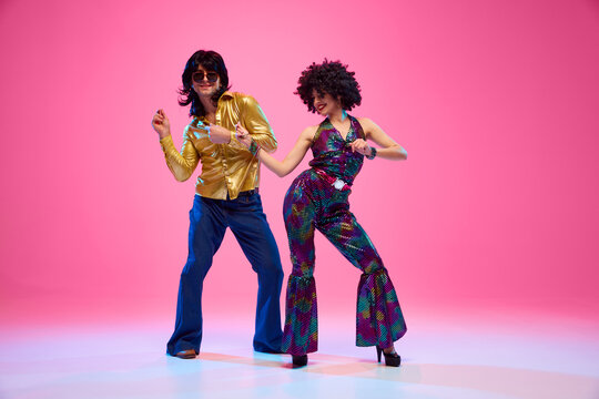Groovy dance couple in bright attire performing disco moves in motion against gradient pink studio background. Concept of American culture, 1970s, 1980s fashion, music, comparisons of eras.