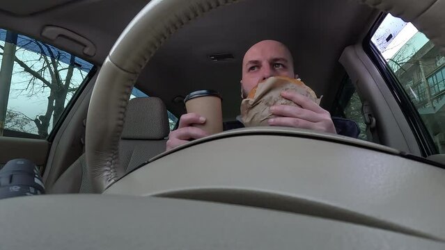 Male driver drinking coffee and eating a burger while driving a car, low angle. The concept of fast food delivery and dangerous driving