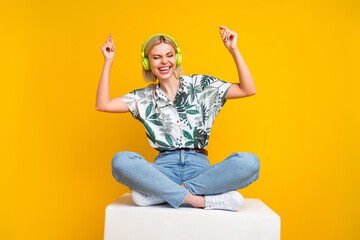 Full size photo of overjoyed girl wear shirt sitting on platform listen uplifting music in headphones isolated on yellow color background