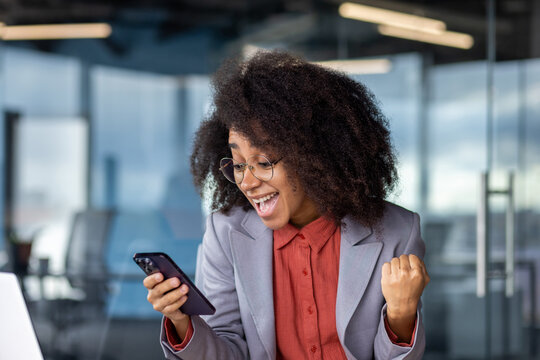 Excited Hispanic Office Manager Raising Hand With Clenched Fist While Looking At Smartphone Screen Indoor. Ecstatic Worker Making Yes Sign With Arm While Getting Message About Unexpected Day Off