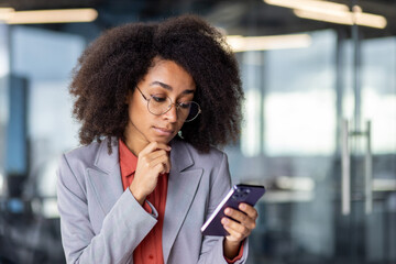 Thoughtful latino woman in grey suit holding chin with hand while looking at smartphone screen indoors. Doubting office worker getting text message with business offer and pondering about answer