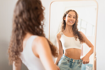 Fototapeta premium teen girl looks at mirror satisfied with weight loss indoor