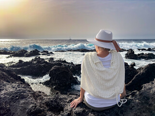 Back view of senior woman sitting on rocky beach watching ocean waves crashing with white foam....