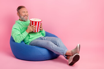 Full length photo of handsome senior man sit bean bag eat popcorn watch film wear trendy green...