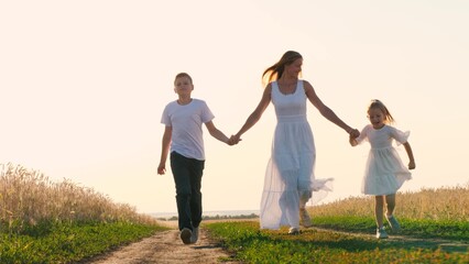 Naklejka na ściany i meble Happy family mother with kids holding hands running on dirt road at sunny field enjoy freedom together. Smiling woman with teen boy and girl kid enjoy freedom outdoor leisure having fun sunset sunrise