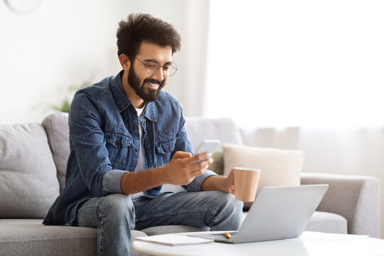 Content indian man browsing smartphone and holding mug of coffee at home - Powered by Adobe