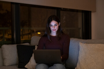 One young woman using laptop by night at home sitting on the sofa searching on web and working...