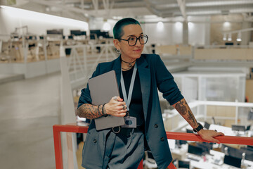 Pretty tattooed female manager in glasses with laptop standing on modern coworking background