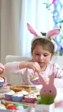 Young mother and her cute little daughter wearing funny rabbit ears are cooking Easter cupcakes sitting at a festive table with basket, eggs and Bunny