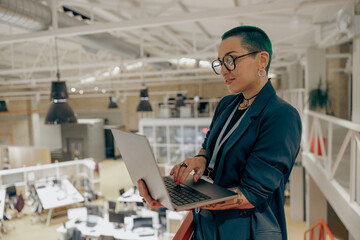 Smiling tattooed business woman in glasses working on laptop while standing on office background