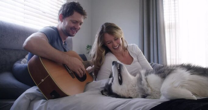 Couple, home and playing guitar with puppy for love and bonding, happy in bed with pet. People with animal, husky dog and musical instrument, time together with trust, commitment and creativity