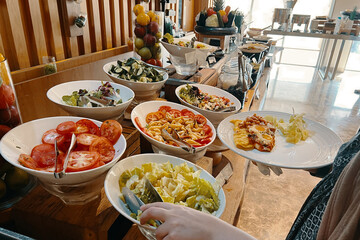 A woman at the buffet table stuffs her plate with food. Cabbage, tomatoes, pasta, cucumbers.