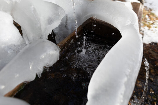 vista in primo piano di gocce d'acqua, che cadono all'interno di una grande bacinella in legno, da un rubinetto all'aperto, in montagna, coperto e circondato da ghiaccio e neve, in inverno