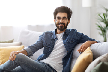 Portrait Of Smiling Young Indian Man Posing On Couch At Home