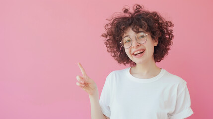 An exuberant young woman with curly hair and glasses, pointing her finger to the side with a wide, joyful smile on a pink background.