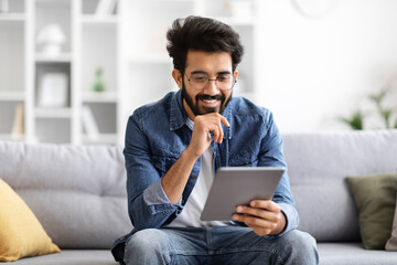 Focused indian man wearing eyeglasses using digital tablet at home