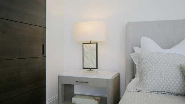 Corner of cozy hotel bedroom with double bed covered with blanket and cushions next to simple nightstand with turned on lamp and dark wooden door aside