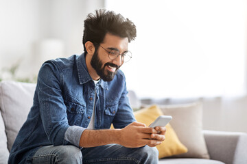 Cheerful bearded indian man looking at his smartphone with smile at home