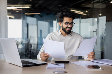 Satisfied Indian young man businessman working with documents, sitting in the office at the table...