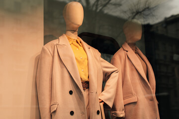 Mannequins in a store window dressed in seasonal fashion. Beige coat and a bright yellow shirt.