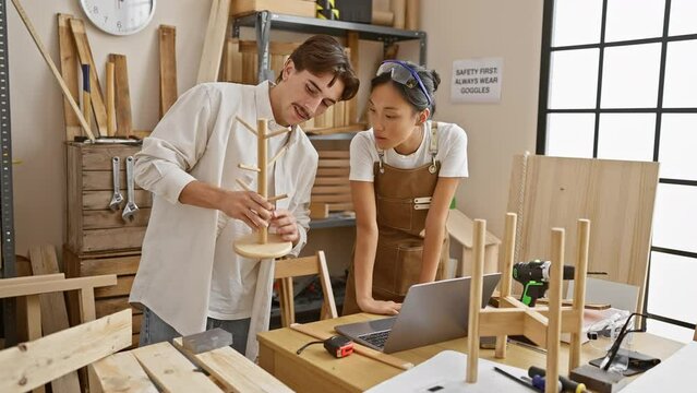 Man and woman collaborate in a bright woodworking workshop, examining handmade furniture by a laptop.