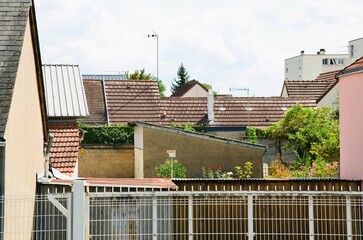 Urban landscape: in the foreground, a metal gate then roofs of houses in red tiles, slates and sheet metal, gardens and in the background white buildings, perspective view with a cloudy sky.