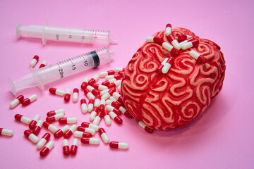 Red human brain with two syringes and a pile of red and white pills and capsules on a pink table.