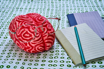 Red rubber brain with glasses and some books next to it on a white surface with green circles.