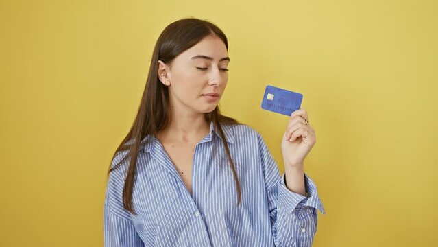 Young, confident hispanic woman deliberating financial moves, credit card in hand. beautiful, self-assured expression; isolated on yellow background, deep in thought.