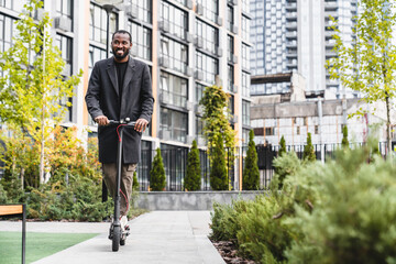 Full length photo of attractive 30s african man riding e-scooter in city park against urban landscape. Businessman manager ceo using ecologically sound clean transport outdoors