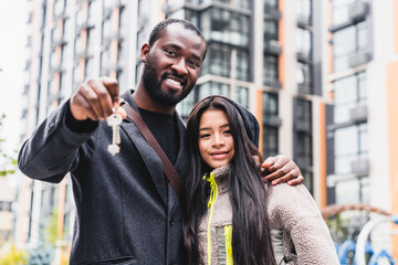 Cheerful happy multiethnic couple holding a key of their new flat against modern block of flats....