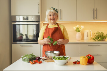 Obraz na płótnie Canvas Senior 60s blond Caucasian smiling woman holding a knife and fresh spinach whole preparing a bowl of healthy diet salad in her rustic eco kitchen. High quality photo