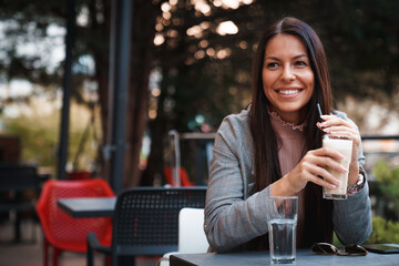 Young attractive woman, sitting by table outdoors, drinking her hot chocolate and smiling to...