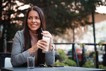 Beautiful young woman sitting in a coffee shop terrace, holding her drink with straw and looking at...