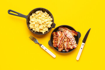 Frying pans of tasty scrambled eggs and bacon on yellow background