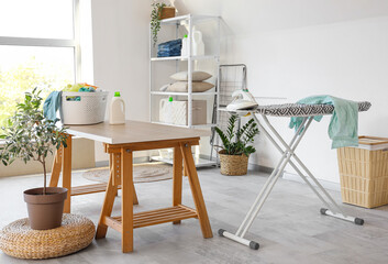 Interior of laundry room with ironing board, clothes in basket and houseplant