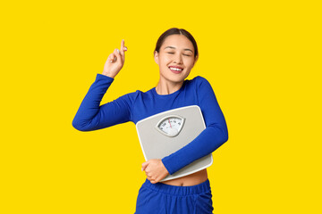 Young woman in sportswear and with weight scales crossing fingers on yellow background. Slimming...