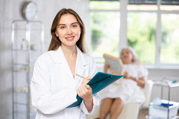 Positive young lady wearing medical coat with pen and folder in her hands standing at treatment room aesthetic clinic