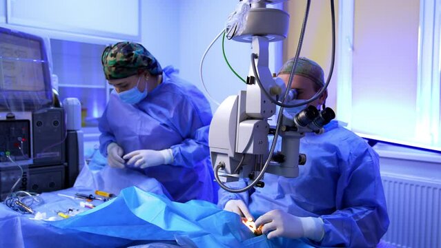 Mid-aged mal surgeon sitting behind the microscope performs ophthalmic operation. Female nurse arranges instruments and materials on the operational stand.
