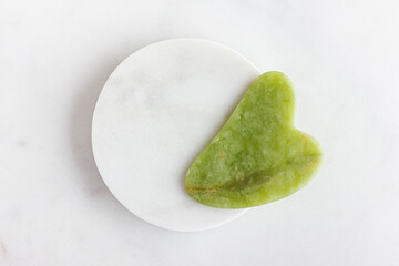 Green jade gua sha massager for face on white marble.