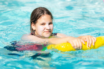 Happy teenager girl with yellow inflatable pillow in swimming pool with blue water on warm summer day on tropical vacations. Summertime activities concept. Cute child girl sunbathing on air mattress