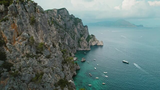 Cliffs on shores of Capri with boats on calm sea waters. Rugged coast of Italian Island in summer day. Aerial view of nature landscape.