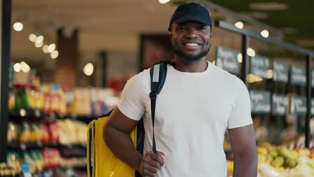Portrait of a happy male delivery worker with Black skin color in a black hat and a white T-shirt who carries a large yellow bag on his shoulders while searching for the necessary goods in a grocery
